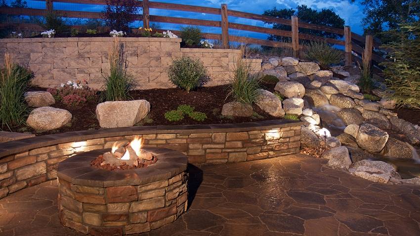 Stone fire pit on a patio with retaining walls and a water feature at dusk
