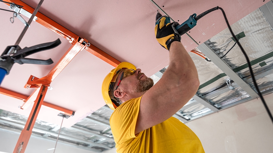 Worker installing drywall on a ceiling using power tools and support equipment.