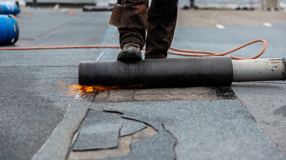 Worker repairing a flat roof using a torch-on felt method with modified bitumen.