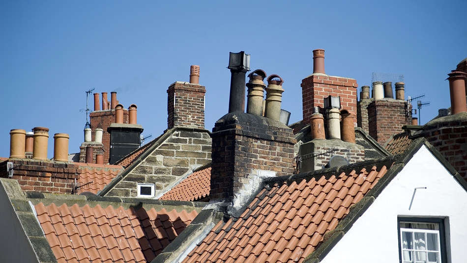 Rooftops of houses with multiple brick and stone chimney stacks topped with various chimney pots under a clear blue sky.