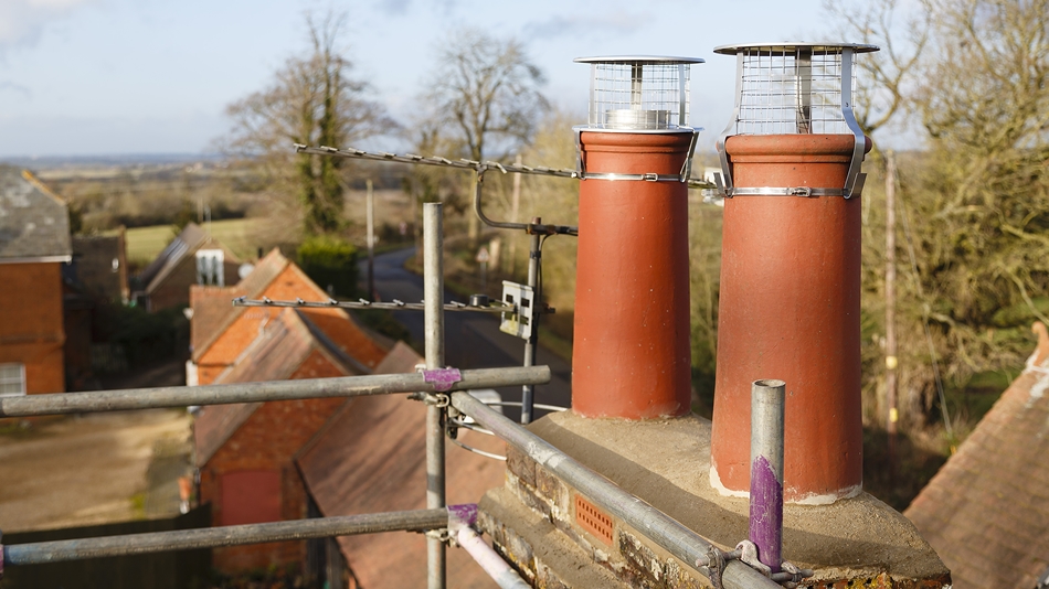 New red clay chimney pots with stainless steel ventilated cowls and fresh flaunching viewed from scaffolding above a rural property.