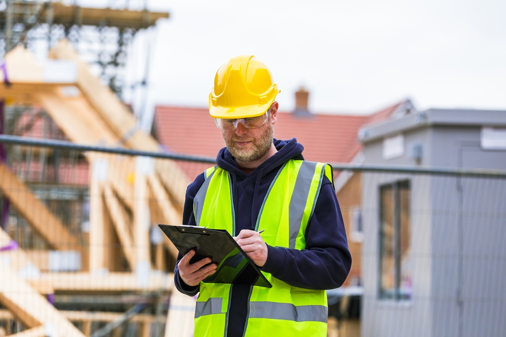 Construction site supervisor in high visibility vest and hard hat completing a checklist on a clipboard.