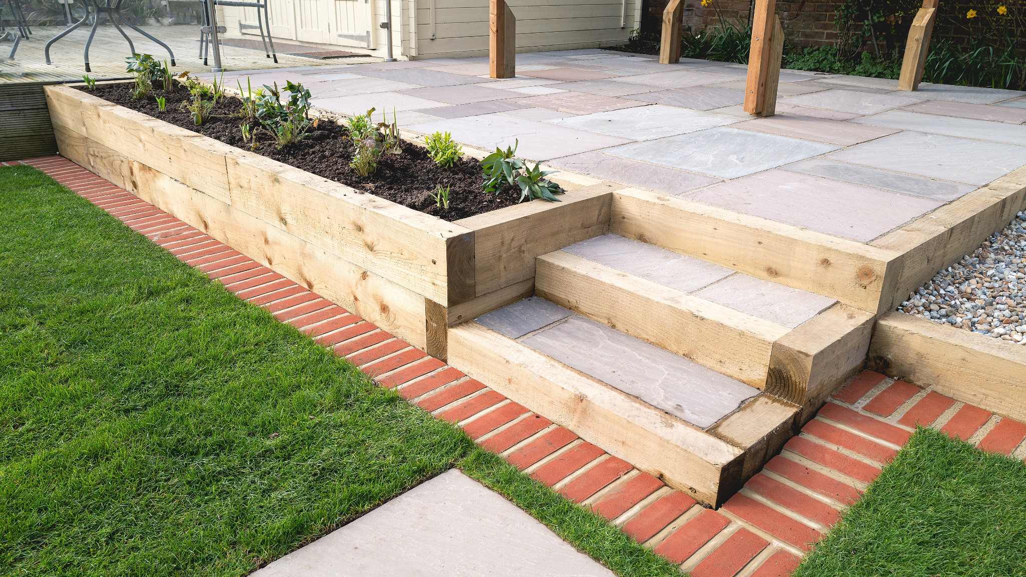 Raised paved patio with timber retaining walls, garden steps, lawn and planting